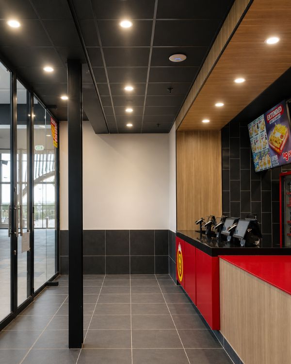 Completed restaurant interior fit-out showing tiled flooring, ceiling lighting, timber wall finishes, black tiled counter area, and red service counter detailing.